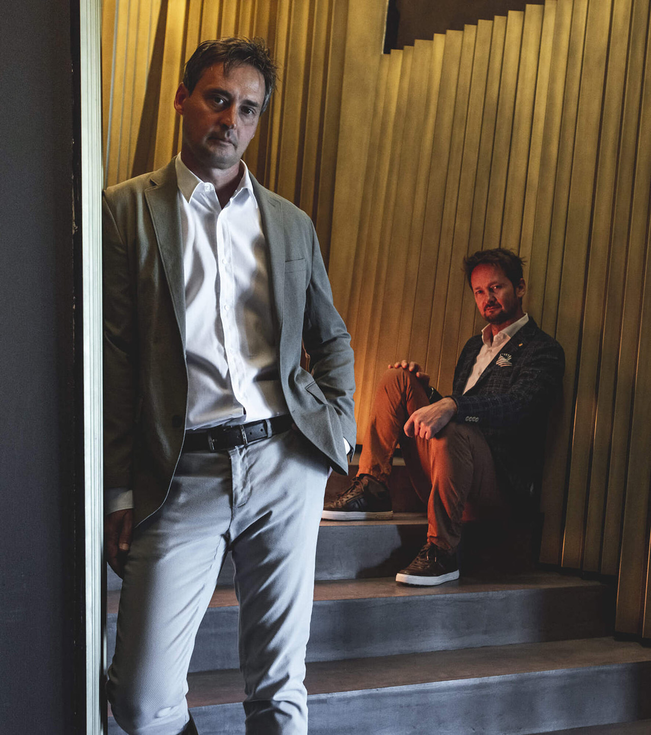 Two men wearing suits, one sitting on the stairs the other learning against a wall.