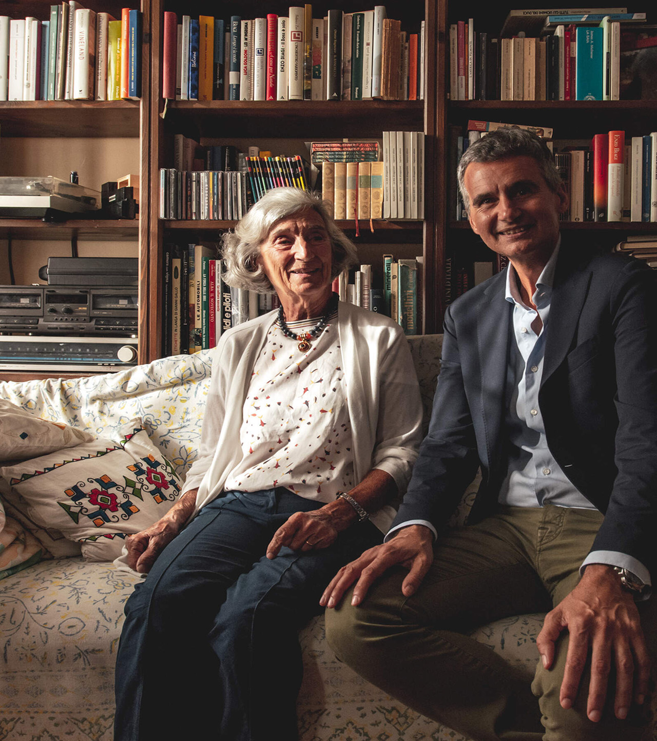 A man and elderly lady sitting on a sofa in front of a bookshelf filled with books.