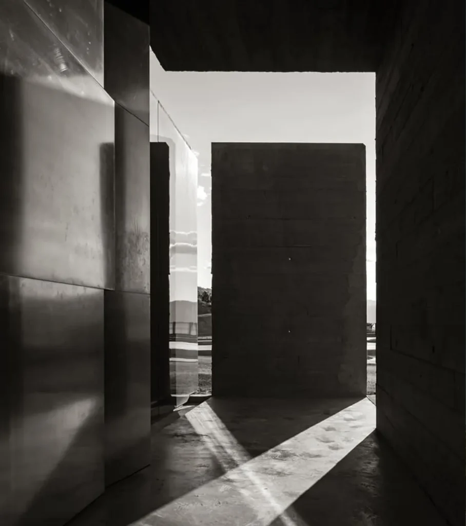 Black-and-white architectural photograph of a modern concrete passageway with dramatic sunlight and geometric shadows.