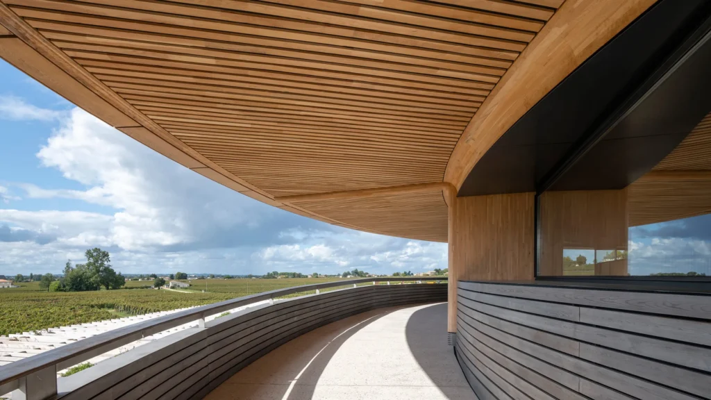 Curved wooden balcony and overhanging roof of a modern winery overlooking expansive vineyards under a partly cloudy sky.