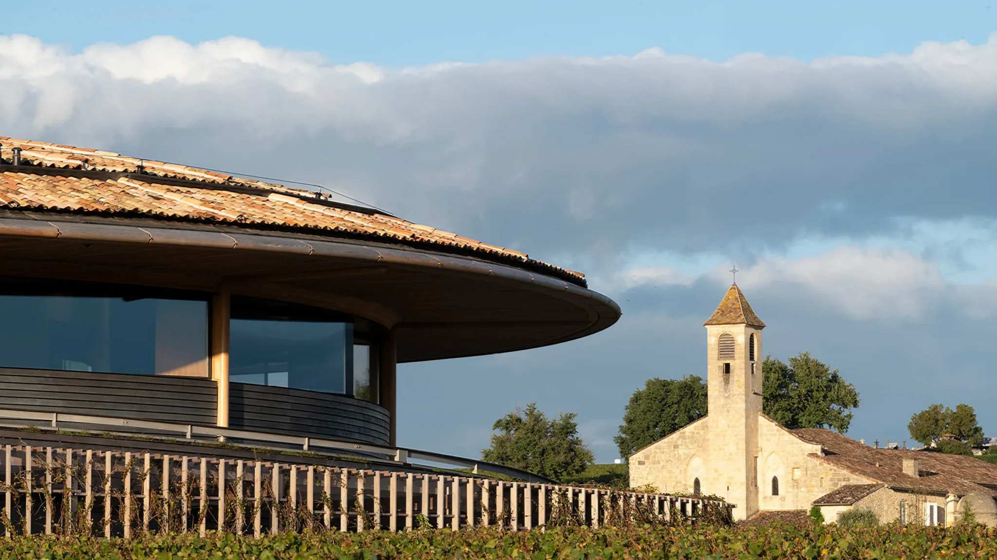 Modern winery building with a curved tiled roof overlooking vineyards and a historic stone church under a cloudy sky.