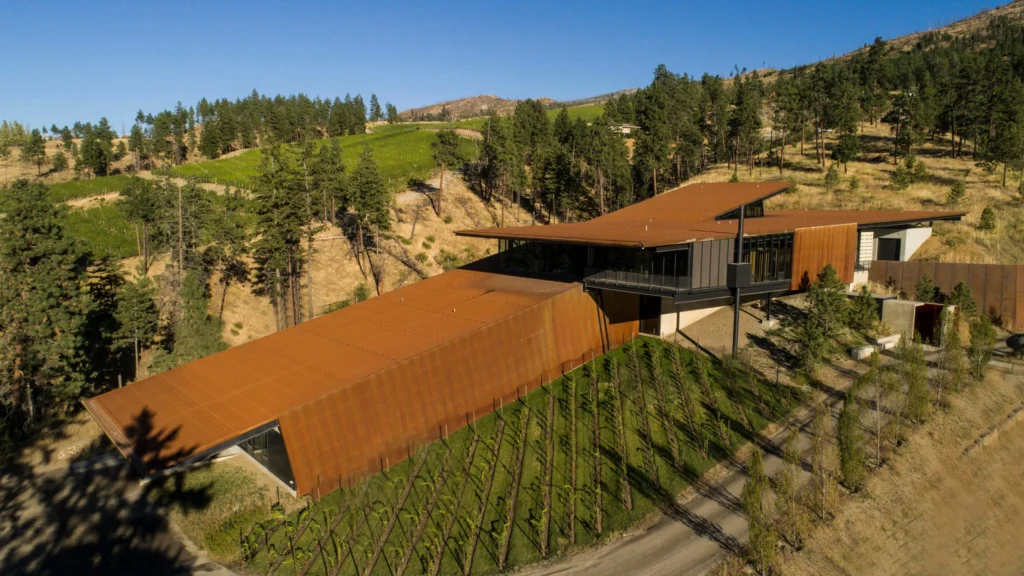 Aerial view of a modern winery with rust-colored roofs, surrounded by vineyards, trees, and rolling hills.