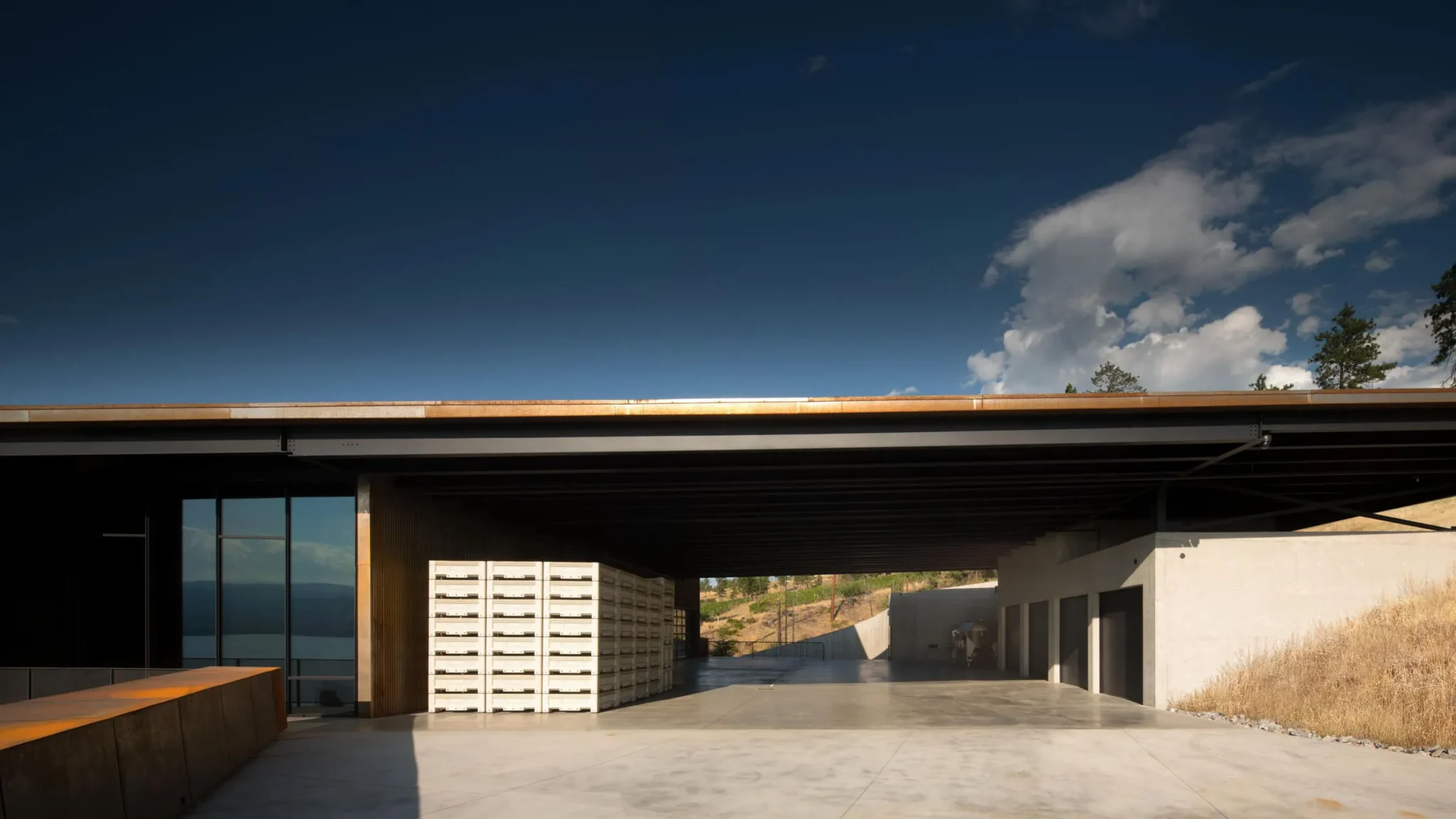 Modern winery exterior with a wide overhanging roof, concrete courtyard, and stacked storage crates beneath a blue sky.