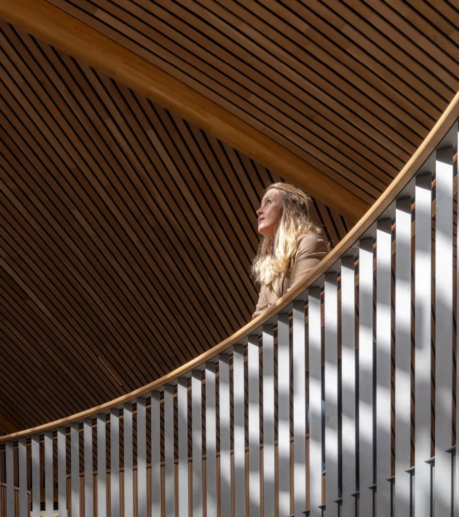 Woman standing behind a curved metal railing beneath a striking wooden slatted ceiling in a modern winery.