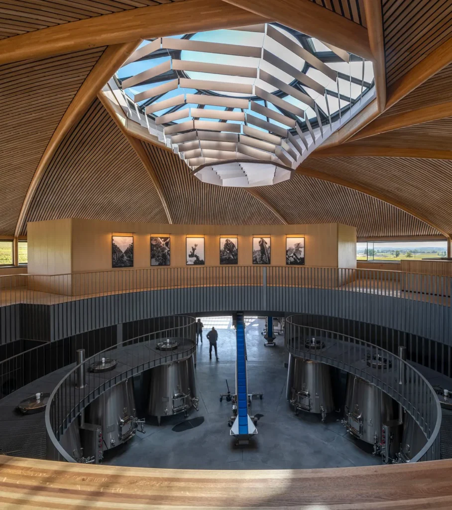 Modern winery interior with a curved wooden ceiling, geometric skylight, and stainless steel wine tanks below.