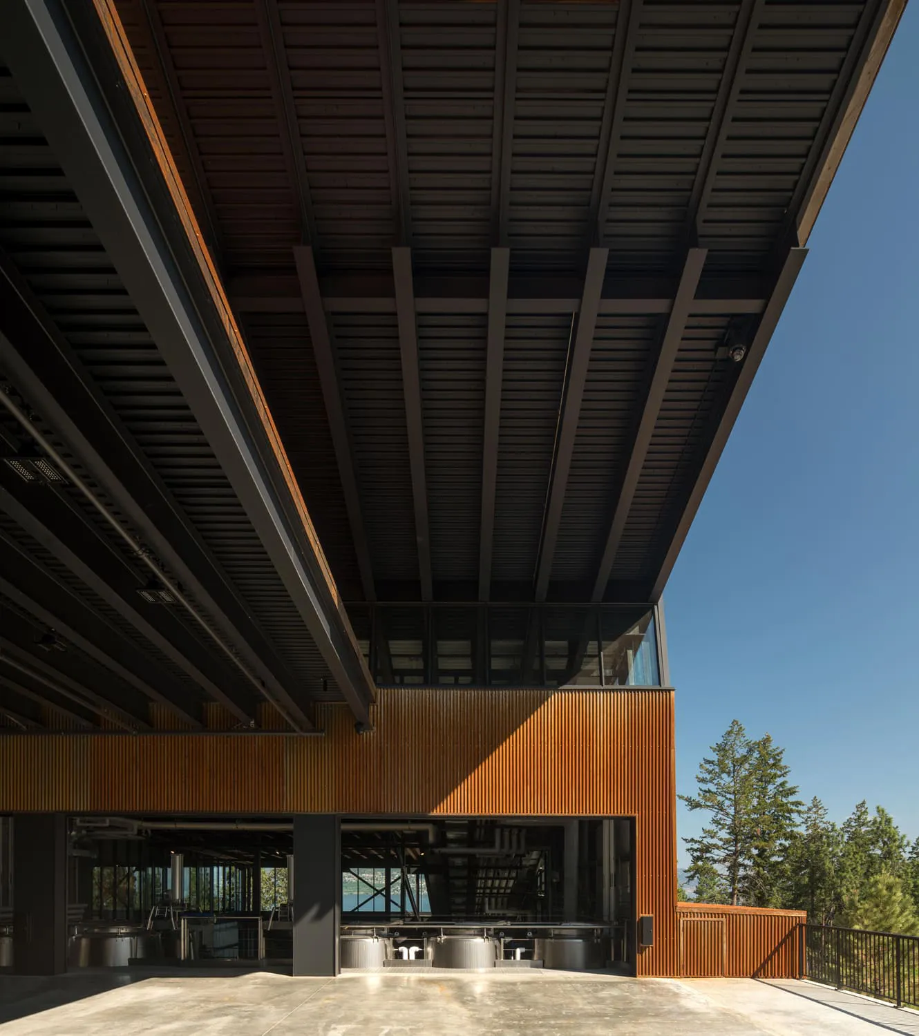 Modern winery building with a large steel roof, wood-paneled exterior, and glass walls revealing stainless steel wine tanks.