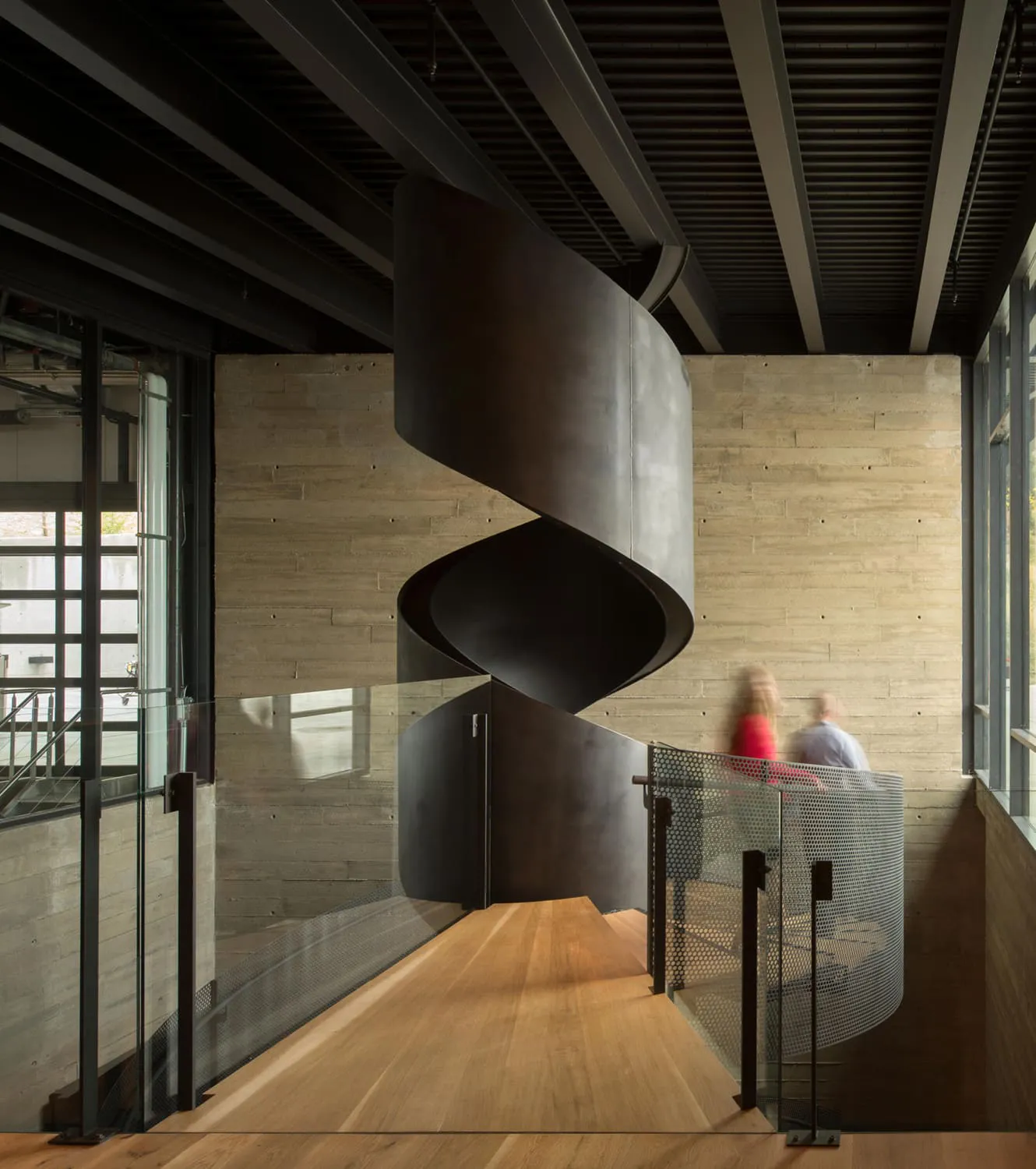 Modern interior featuring a sculptural black spiral staircase, concrete walls, wooden flooring, and glass railings.