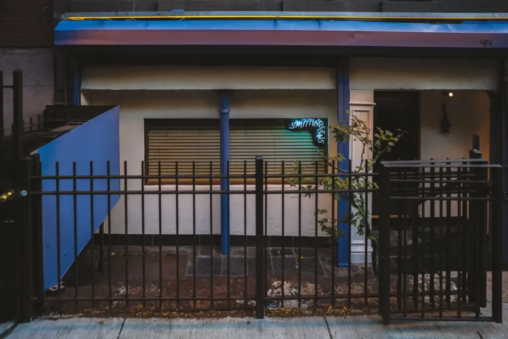 Exterior of a small restaurant entrance with blue accents, black fencing, and a glowing neon sign.