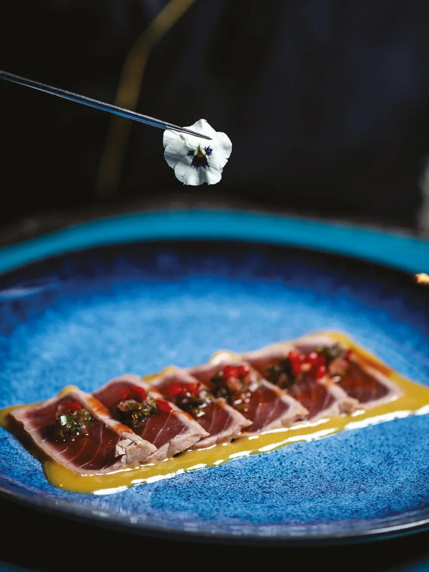 Seared tuna tataki on a blue plate, garnished with a flower held by tweezers