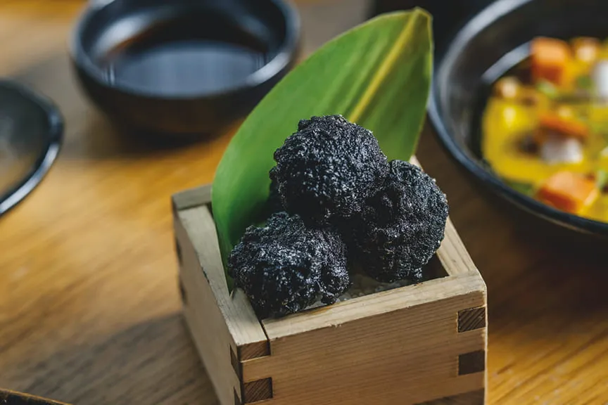 hree black crispy bites served in a wooden box with a green leaf.
