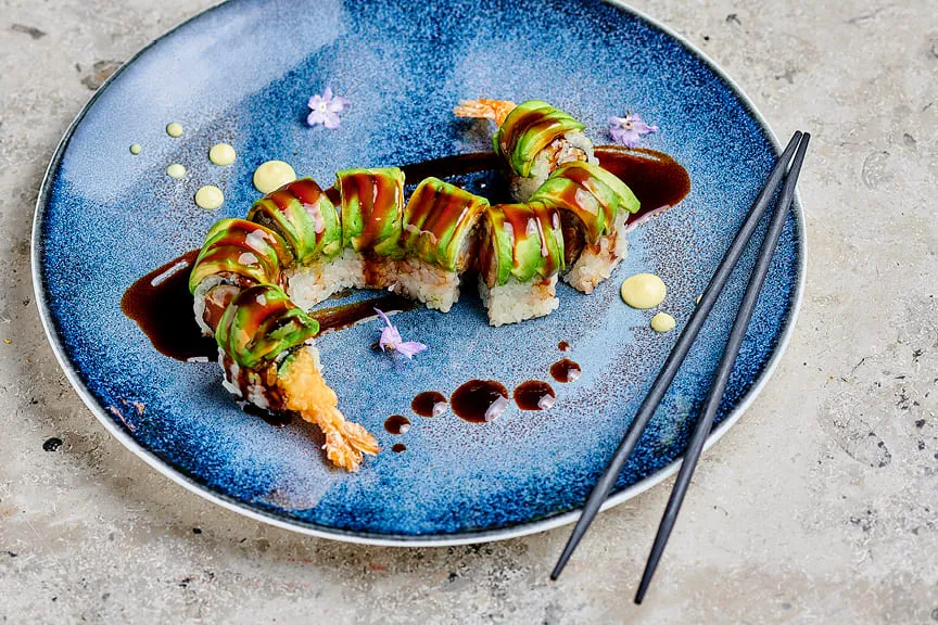 Avocado-topped sushi roll with shrimp, sauces, and edible flowers on a blue plate.