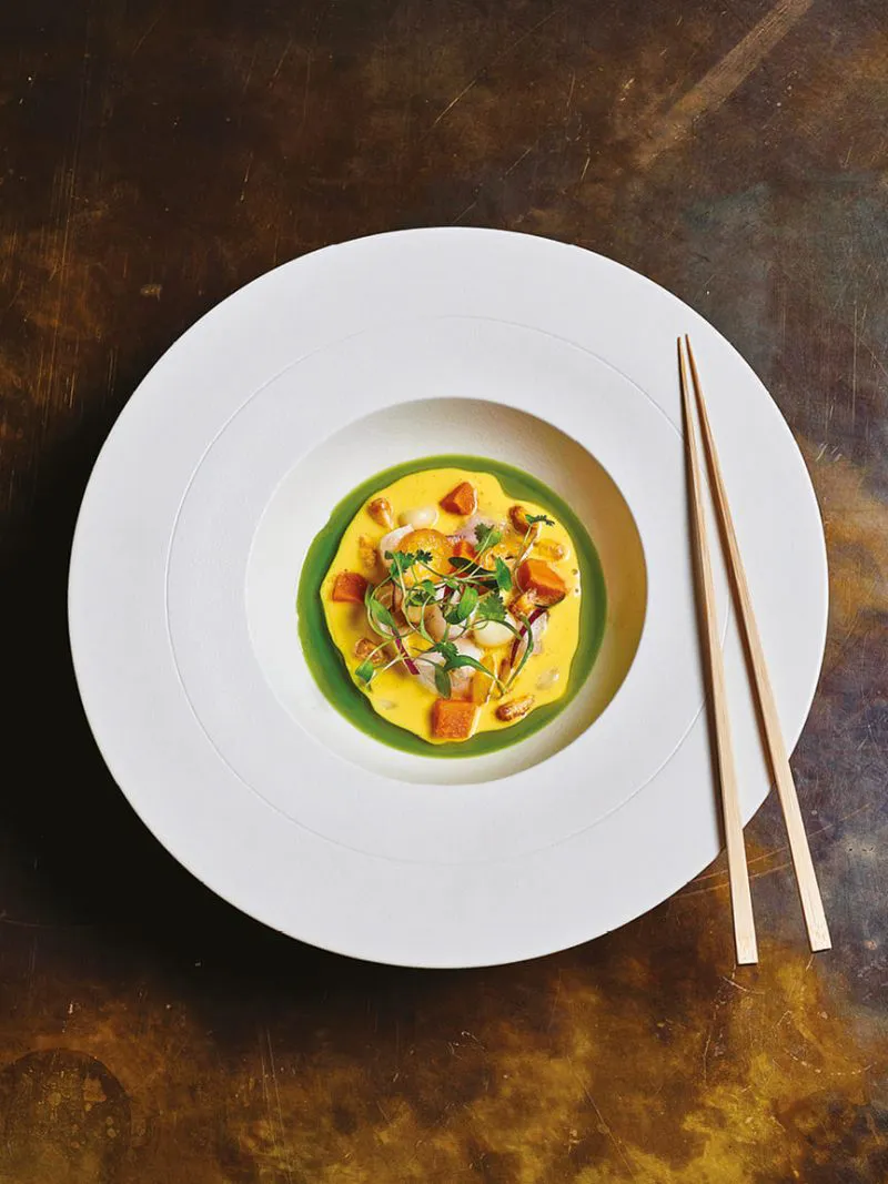 Artfully plated dish with yellow and green sauces, garnished with herbs, served with chopsticks.