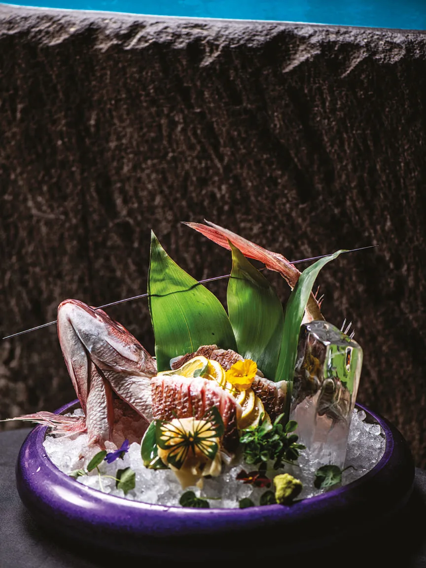 Sashimi platter on ice with edible flowers and leaves in a purple bowl