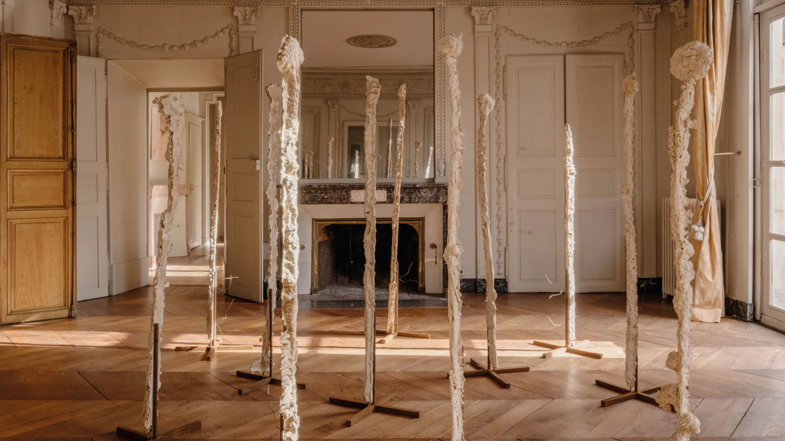 Tall, textured white sculptures arranged in an elegant room with ornate walls, a fireplace, and wooden floors.