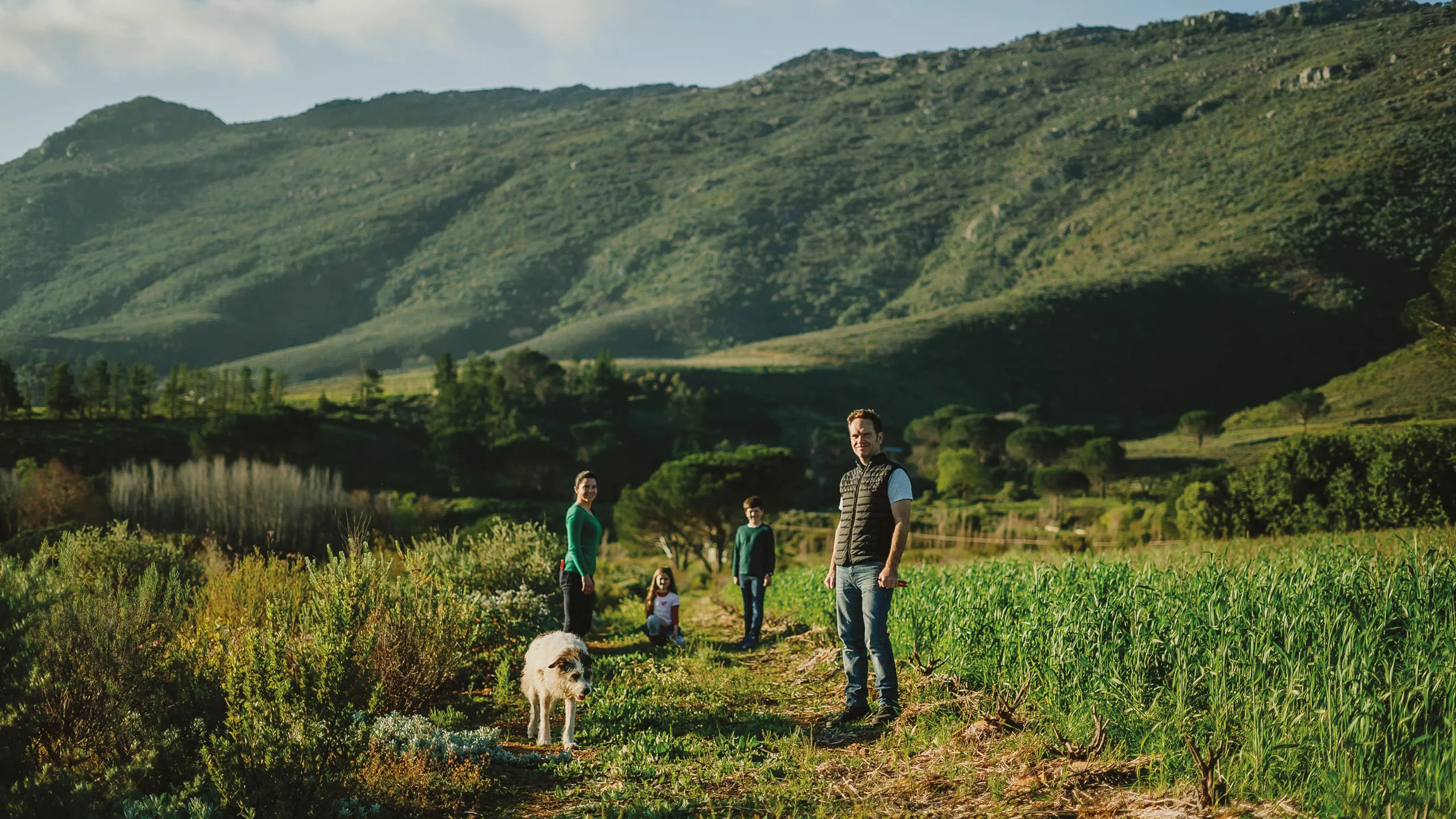 Family with a dog standing among green crops in a scenic mountain valley.