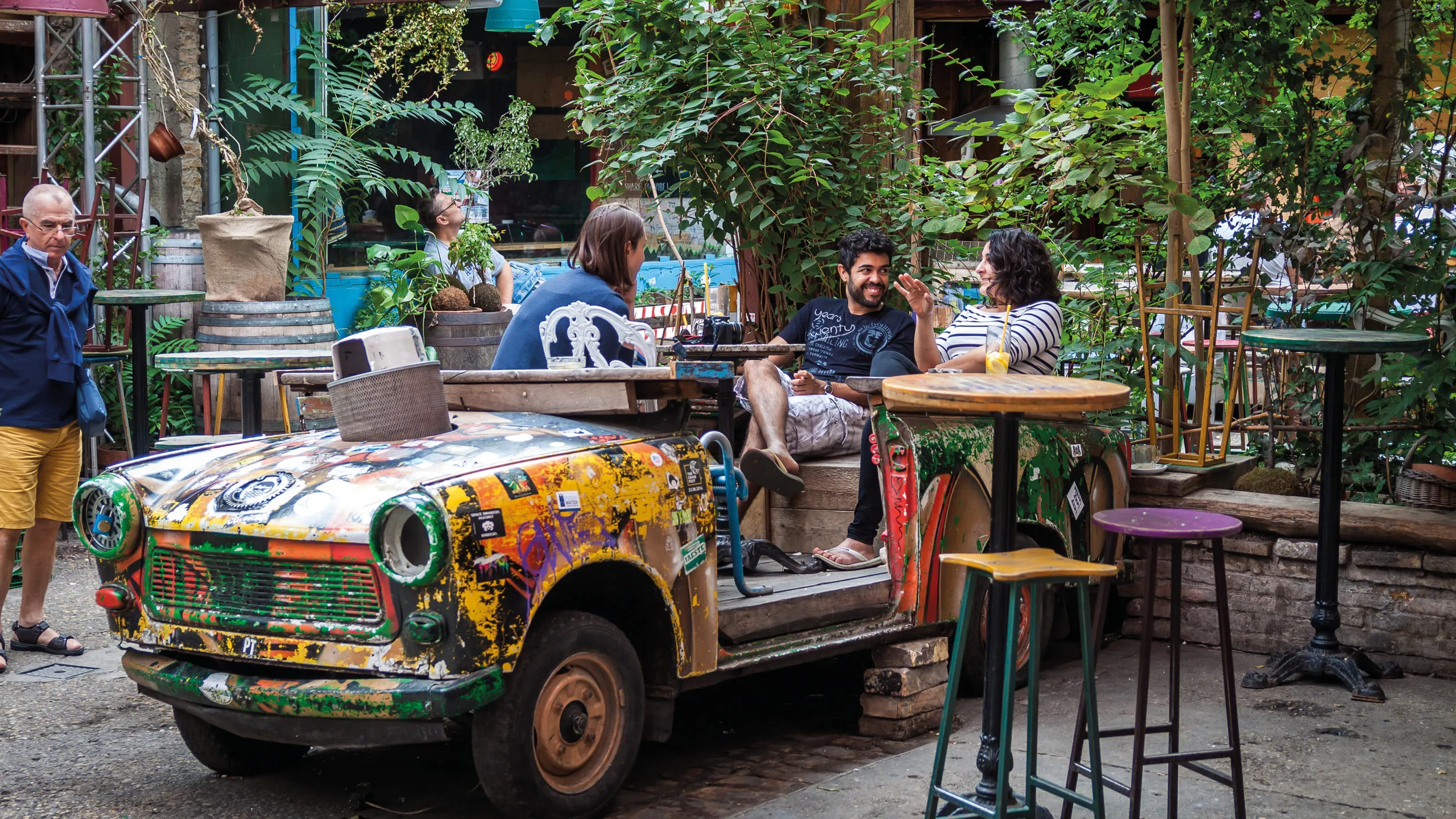 People relaxing at an outdoor café with lush greenery and colorful vintage car seating.