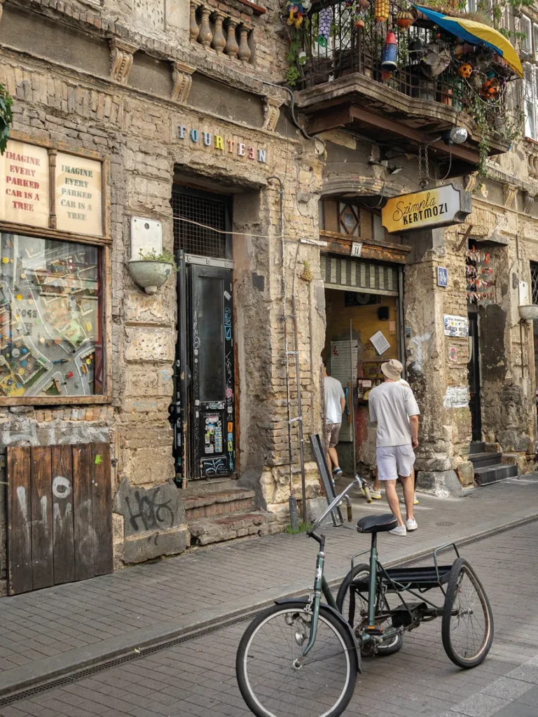 Rustic brick building with a decorated balcony, café entrance, and a vintage three-wheeled bicycle parked outside.