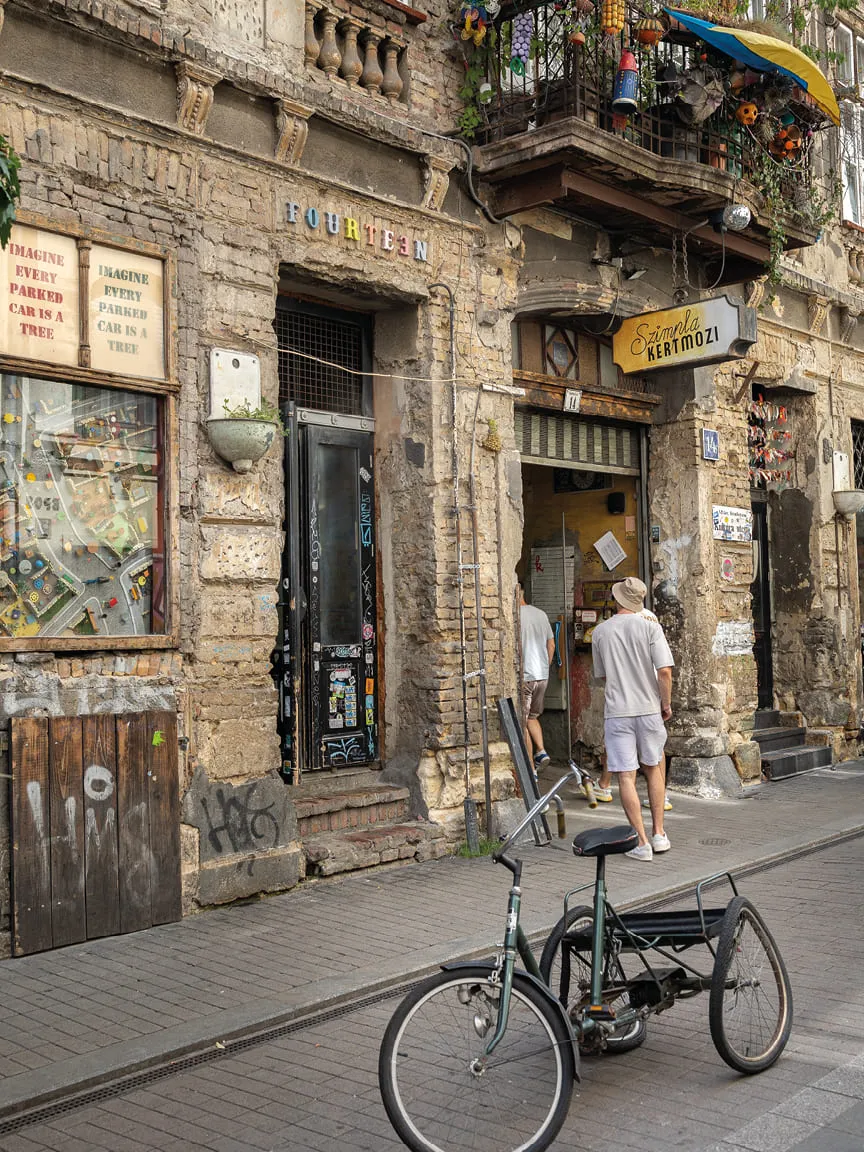 Rustic brick building with a decorated balcony, café entrance, and a vintage three-wheeled bicycle parked outside.