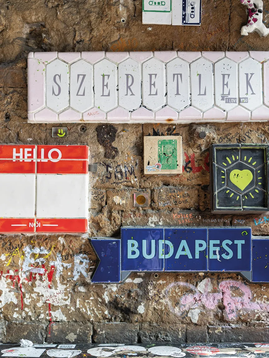 Colorful tiled signs reading “Szeretlek,” “Hello,” and “Budapest” on a graffiti-covered stone wall.