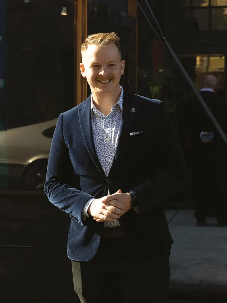 Smiling man in a suit standing outside a restaurant.