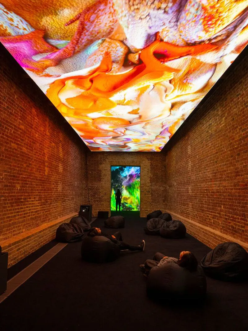 Immersive digital art installation with colorful ceiling projections and visitors relaxing on beanbags in a brick-walled gallery.