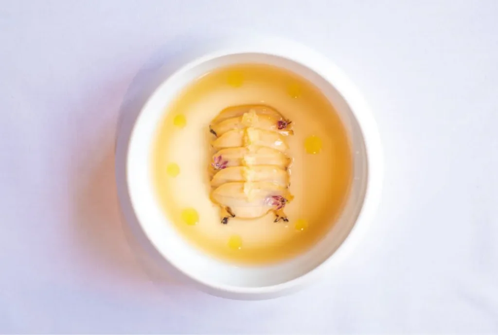 Delicately sliced seafood arranged in a golden broth with small drops of oil and floral garnishes.