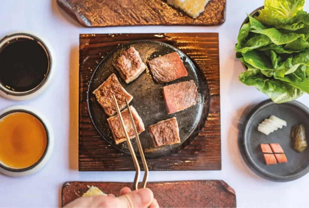 Slices of seared beef served on a hot plate with dipping sauces, lettuce, and pickled vegetables.