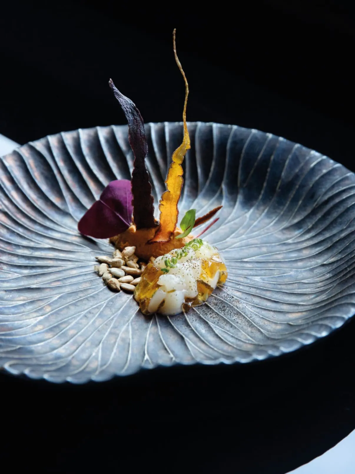 Artfully plated fine-dining dish with colorful vegetables and delicate garnishes on a textured grey plate.