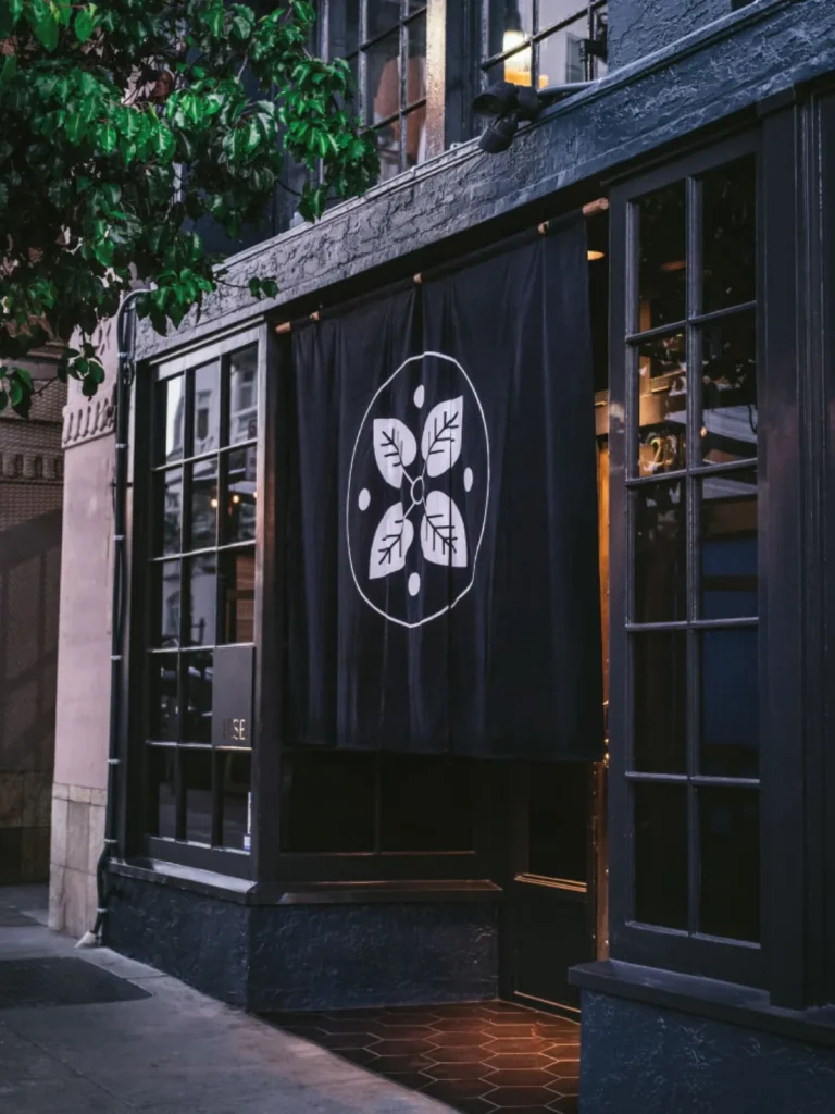 Dark restaurant exterior with large windows and a black curtain featuring a white floral logo.